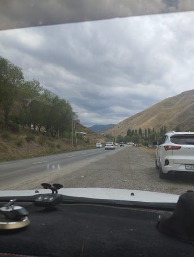 The image shows a cloudy day on a highway in Karangisay, Uzbekistan. The road is paved with a gravel shoulder on the right side. Several cars are visible on the highway. On the left side of the road, there is a gentle slope covered with dry grass and sparse trees. Further up the slope, a line of trees with lush green foliage is visible, with a few small houses nestled among them. To the right of the highway, a large, barren, yellowish-brown mountain dominates the background. A white SUV is parked on the gravel shoulder in the foreground, facing away from the camera. The vehicle's license plate is visible and reads "85 J B". In the foreground, inside what appears to be a vehicle, a dashcam is mounted on the windshield.