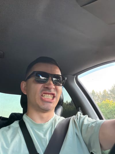 A man wearing sunglasses and a light green t-shirt is in the driver's seat of a car, with his mouth open and teeth bared in a grimace. The interior of the car, including the dashboard and a seatbelt, is visible. Outside the car, through the window, trees and a bright sky can be seen. The image was taken in Kamyennoye, Russia.