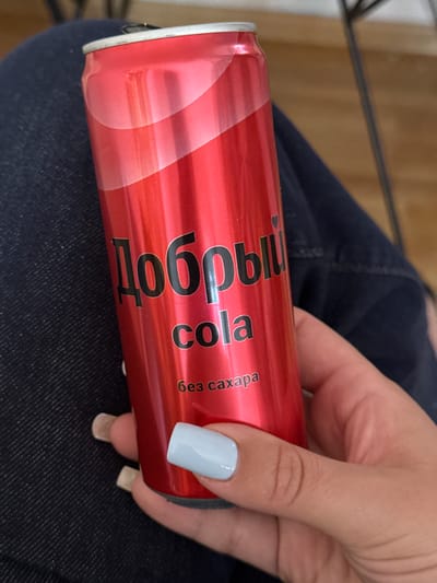 A person's hand with light blue nail polish is holding a red aluminum can of soda. The can has black text on it, which appears to be in Cyrillic script. The text reads "Добрый cola" vertically, with "без сахара" below it. The can is ribbed and has a metallic sheen. The person is wearing dark denim pants, which are visible in the background. The lighting suggests it is daytime, but there are no strong shadows or direct sunlight. The scene appears to be indoors or in a shaded outdoor area. The focus is on the hand and the can, with the background out of focus. The overall impression is a casual, everyday moment.