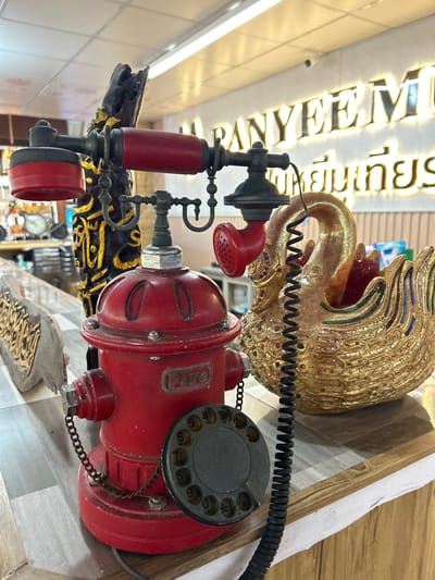 A close-up, eye-level shot captures a vintage red fire hydrant-shaped rotary telephone and a golden swan figurine on a wooden counter in Tambon Ko Panyi, Thailand. The telephone, with its red earpiece and receiver, features a black coiled cord and a rotary dial with numbers. A decorative, carved black and gold column stands behind the telephone. To the right of the telephone is the ornate golden swan, adorned with colorful mosaic details, positioned in front of a white wall with illuminated signage in English and Thai. The background shows blurred elements of a shop interior, including shelves and lighting fixtures. The counter surface is made of patterned wood-look material.