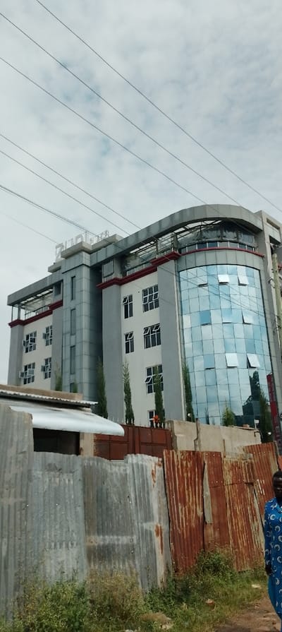 The image depicts a multi-story building and its immediate surroundings in South Kasipul ward, Kenya, under an overcast sky.

The prominent subject is a modern, five-story building constructed with grey and white facades, featuring a large, curved blue-tinted glass section on its right side. Several windows on both the glass and solid sections of the building are open. The word "ZUPAN" is visible in white letters atop the building's central roofline. Red trim accents are present beneath some window sills and along roof edges. Tall, slender evergreen trees are planted in front of the building's ground level, partially obscured by a dark brown gate.

In the foreground, a fence constructed from corrugated metal sheets extends across the lower portion of the frame. The left section of the fence is composed of silver-grey galvanized sheets, while the right section consists of heavily rusted, dark orange-brown sheets. Behind the left part of this fence, a small, low-rise structure with a light-colored, sloped roof is visible. Sparse green and dry weeds and grass grow along the base of the fence.

In the lower right corner, a person is partially visible, facing slightly away from the camera but with their head turned towards the left. They are wearing a long-sleeved, blue garment with a repeating white pattern.

Above the building, multiple parallel power lines traverse the frame diagonally from the upper left to the lower right. The sky is uniformly overcast with visible cloud formations.