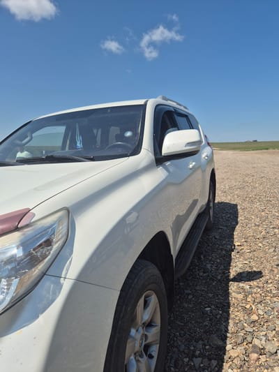 A white SUV is parked on a gravel road with a wide, open grassy plain in the background. The sky is bright blue with a few scattered white clouds. The car is angled towards the viewer from the front left, showing its headlight, hood, windshield, side mirror, and part of the passenger door and running board. The terrain suggests a rural or undeveloped area. There is a person visible inside the car through the windshield. In the distance, on the horizon, there are some structures that appear to be buildings or other man-made objects, possibly associated with agriculture or rural living, in the vicinity of Novo-Ishim Rural District, Kazakhstan.