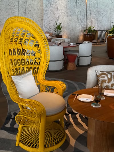 The image shows an indoor seating area with a focus on a bright yellow wicker chair. The chair is ornate, with intricate weaving patterns and a fan-shaped back. A white pillow rests on the seat of the chair. To the left of the yellow chair, there are two cylindrical white ottomans with gray straps. Behind the ottomans, there is a large, textured wall and a round wooden table set with plates, glasses, and chopsticks. A patterned rug with gray and white stripes is visible on the floor. In the background, a potted plant is placed on a shelf, and part of a light-colored couch with a patterned cushion is visible. The overall scene suggests a contemporary, possibly high-end, dining or lounge area. There are no people visible in the image, and the lighting suggests it is daytime, likely indoors with artificial lighting. The location is unknown, but is described as potentially being the Maldives. There is no visible text in the image.