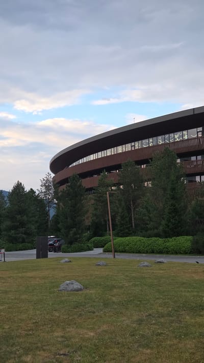 The image captures a modern, curved building with a brown, slatted facade and large windows. The windows have intricate, illuminated designs visible from the outside. In front of the building, there is a well-maintained lawn with several large rocks scattered across it. A paved walkway leads to the building, with a few vehicles and two people visible near a dark structure on the left side of the walkway. Tall evergreen trees and lush green bushes surround the building and the lawn, suggesting a well-landscaped environment. The sky above is partly cloudy, with hues of blue and gray, indicating it might be late afternoon or early evening. The scene appears to be located in a serene, possibly suburban or resort-like setting. The specific location in Озерное (майминский), Russia, is not discernible from the visual information alone.