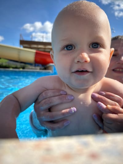 A baby with blue eyes is being held in a swimming pool by an adult whose hands are visible. The baby's head is close to the camera, showing a portion of their face. In the background, a water slide and blue sky with clouds are visible. The setting appears to be an outdoor swimming pool, likely in an area with recreational facilities. The city of Юбилейный, Russia is not explicitly depicted in the image, but the presence of a water slide suggests a public or private recreational area.