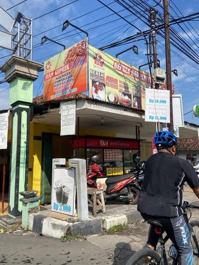 A man wearing a black shirt and blue and black cycling pants stands in front of a food stall in Kecamatan Tulangan, Indonesia (latitude: -7.482, longitude: 112.648). The stall has a red awning and several signs, one that reads "Ayam Geprek Rp 10,000" and another that reads "Bebek Kalasan Rp 20,000," suggesting that the stall sells fried chicken and duck. The man is looking at the camera. He has a bicycle parked next to him and there are several other bicycles in the background. The street is paved and there are power lines overhead. The weather is sunny and the sky is blue. The image is taken at a low angle.