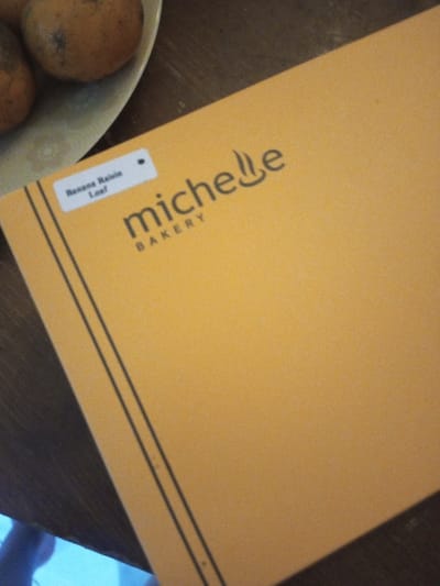 An orange bakery box sits on a wooden surface. The box, labeled "Michelle Bakery," has a white sticker in the upper left corner reading "Banana Raisin Loaf." Two thin, dark lines run down the left side of the box. A partially visible plate with two oranges rests in the upper left corner of the image. A blue object is faintly visible in the bottom left corner.