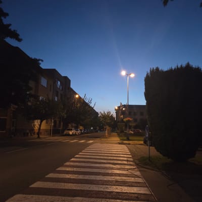 This media file depicts a street scene at dusk in Piešťany, Slovakia. The sky is a deep, clear blue, transitioning to a lighter hue near the horizon, suggesting the twilight hours. A tall street lamp with two glowing bulbs illuminates the area, casting warm light onto the street.

The foreground is dominated by a pedestrian crossing, its white stripes sharply defined against the darker asphalt. To the left of the crossing, a row of parked cars lines the street, their forms silhouetted against the dim light. Beyond the cars, buildings rise, their windows reflecting the fading light. Trees with dark foliage are interspersed along the street and in front of the buildings, some partially obscuring the structures. A large, dense hedge runs along the right side of the image, its dark form contrasting with the illuminated sky.

A blue circular traffic sign with a white arrow indicating a right turn is visible on a pole near the hedge. The overall atmosphere is serene and quiet, indicative of the end of the day. No people are visible in the frame. The image is oriented vertically, with the sky at the top and the street running diagonally from the bottom left to the top right.