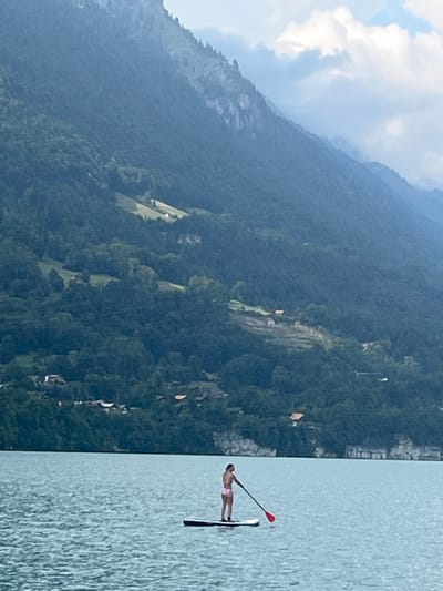 The image depicts a female individual on a stand-up paddleboard on Lake Brienz, located near Bönigen, Switzerland. The person, an adult female, is positioned centrally in the midground, standing upright on a dark-colored paddleboard. She is wearing a light pink two-piece swimsuit and holds a paddle with a red blade, angled to the right. The water surface exhibits minor ripples. In the background, steep, forested mountains rise directly from the lake, heavily covered in dark green vegetation. Scattered across the mountainsides are several small, cleared green pastures, some containing individual white or light-colored structures, likely chalets or farmhouses. Clusters of residential buildings are visible at the base of the mountains along the lakeshore, integrated into the treeline. A distinct rocky cliff face is visible protruding from the forest along the waterline on the right side of the image. The sky above the mountains is partly cloudy, with areas of blue sky interspersed with white and light grey clouds.