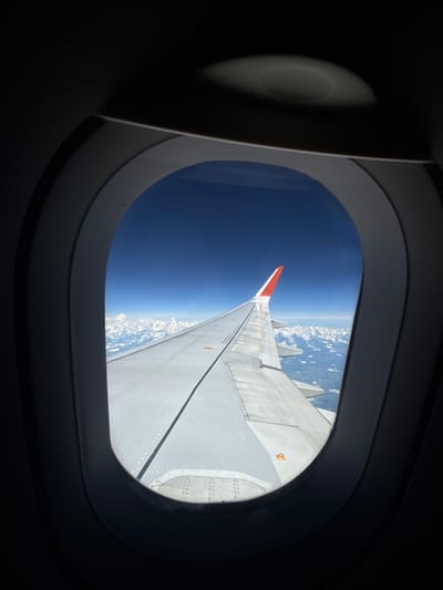 This image is a view from an airplane window, looking out at the wing and clouds. The sky is a deep blue, and the clouds are white and fluffy. The wing is white with a red tip. There are no people or any indication of the city of Усть-Ламенка, Russia. The scene is serene and vast, suggesting a high altitude flight.