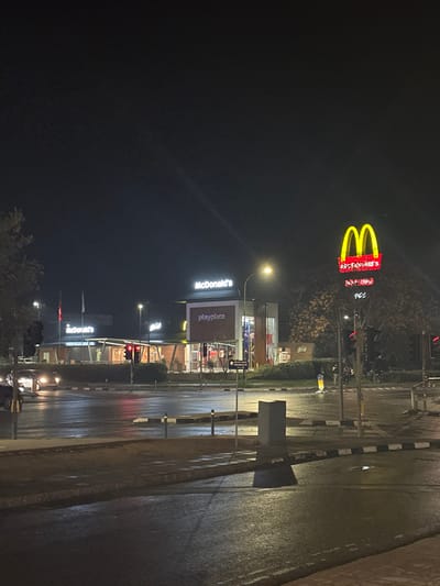 This media file captures a night scene in Engomi, Nicosia, Cyprus, centered around a McDonald's restaurant. The image is oriented sideways, with the ground appearing on the right.

The main subject is a brightly lit McDonald's establishment. A large, iconic golden arches sign with the text "McDonald's" in red is prominent in the upper part of the image, standing tall above the tree line. Below this, the McDonald's restaurant building itself is visible, illuminated from within. Another smaller "McDonald's" sign can be seen on the building. Adjacent to the building is a sign displaying "playplace" on a reddish background.

The scene depicts a wet or damp road, reflecting the artificial lights, suggesting recent rain. Several streetlights cast pools of light on the asphalt. Multiple traffic light poles are present along the road, with some showing illuminated red lights. The road features clear white lane markings and a distinctive black-and-white striped curb or barrier on the right side. Faint outlines of vehicles, possibly with headlights or taillights, are visible in the distance on the road, indicating traffic. Some trees and vegetation are scattered throughout the background and along the roadside. There are no people clearly visible, and no specific event or activity is taking place beyond the general urban night scene.