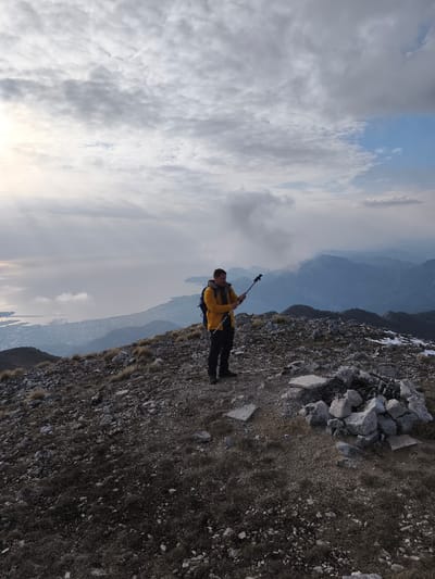 An adult male stands on a rocky mountain ridge in Mali Mikulići, Montenegro. He is positioned slightly right of the frame's center, wearing a bright yellow hooded jacket, dark trousers, and a blue backpack. He holds a black and silver hiking pole horizontally with both hands, appearing to adjust it. The immediate ground is barren, composed of dark gravel, rocks, and sparse patches of dry, brown grass. To his right, a circular arrangement of light-colored rocks and broken concrete slabs forms a ground structure. In the background, a large coastal city borders a body of water, possibly a bay, with several vessels visible. Beyond the city and water, multiple layers of distant mountains are visible, some peaks partially obscured by low-lying clouds or mist. The sky is largely overcast with grey clouds, though a bright area consistent with sunlight is present on the far left horizon. Patches of snow are visible on the higher ground to the far right.