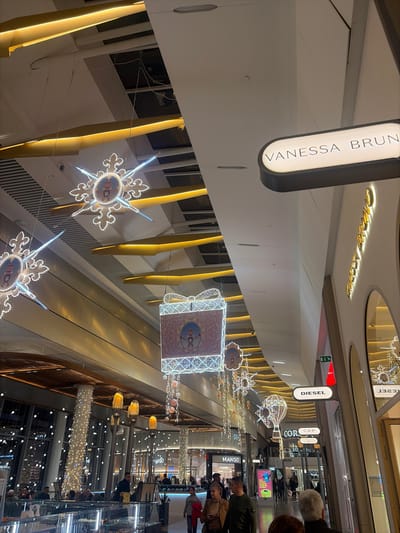 Captured from a tilted angle, the interior showcases a modern shopping mall adorned with holiday lights and décor. Signage for stores like "Vanessa Bruno", "Diesel", and "Mango" is visible. The architecture features a bright, minimalist design with vertical golden accents and reflective surfaces. Patrons casually stroll through the mall, adding life to the scene.