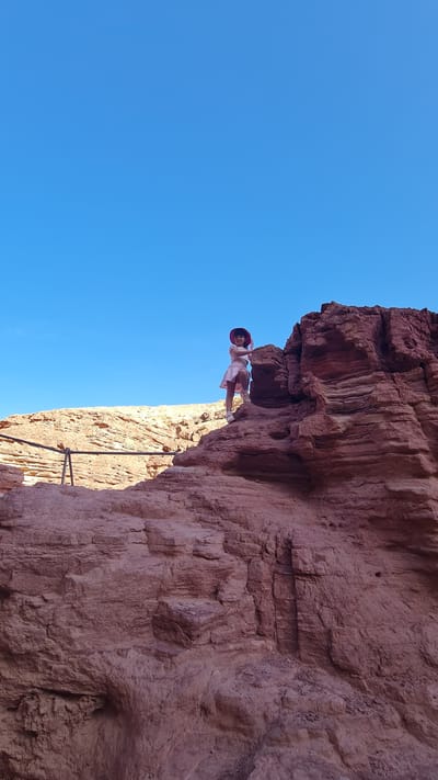 A girl stands on the edge of a red rock formation under a clear blue sky in Hevel Eilot Regional Council, Israel. The rock formation is layered and textured, with a handrail visible below the girl. She is wearing a light-colored dress and a hat.