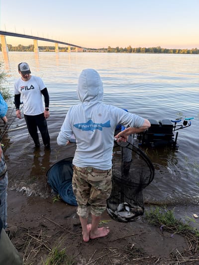 The image shows a group of people by a river, likely engaged in fishing. In the foreground, a person wearing a light blue hooded shirt with a "FINNTRAIL" logo and camouflage pants holds a fishing net filled with fish. To the left, another person in a white "FILA" t-shirt and black pants stands in the water. In the background, a large bridge spans the river, with a treeline visible on the opposite bank. The sun appears to be setting, casting a warm glow over the scene. The location is identified as the city of Kambarka, Russia.