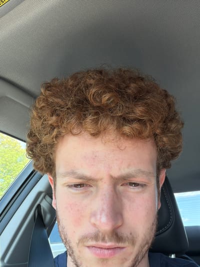 The original image, rotated 90 degrees counter-clockwise, presents a close-up portrait of a light-skinned male individual, from the chest up, inside a vehicle. He has a full head of curly, light brown or auburn hair and light brown eyes that look directly at the camera with a neutral to slightly content expression. He also has light facial hair, including stubble forming a mustache and beard. His complexion shows some freckles and a few small blemishes.
The setting is clearly a car interior. A gray, subtly patterned headliner or ceiling fabric is prominent on the upper left side of the frame. A black car seat headrest is visible behind the person's right ear and head. Through a car window in the lower right, a bright blue sky with some soft clouds and vibrant green foliage indicates a clear, sunny day. The individual is wearing a dark blue garment, possibly a shirt, visible on his right shoulder and chest, with a black seatbelt draped across him. On the far left, a yellow strap with the partial text "VER" is visible, likely part of a safety warning label (e.g., "OVERHEAD AIRBAG" or "WARNING"). The image appears to be a casual selfie taken during daylight hours within a vehicle, possibly in Ridgefield Park, United States of America.