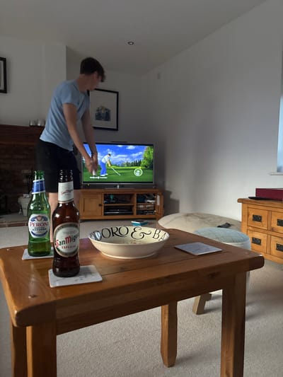 A boy plays a golf video game on a television. The game features a golfer on a green course. In the foreground, two bottles of beer – San Miguel and Peroni – rest on a wooden table alongside a decorative bowl and coasters. The table's design and the surrounding furniture give a sense of being indoors, likely in a living or dining area. The scene appears relaxed, suggesting leisure time.