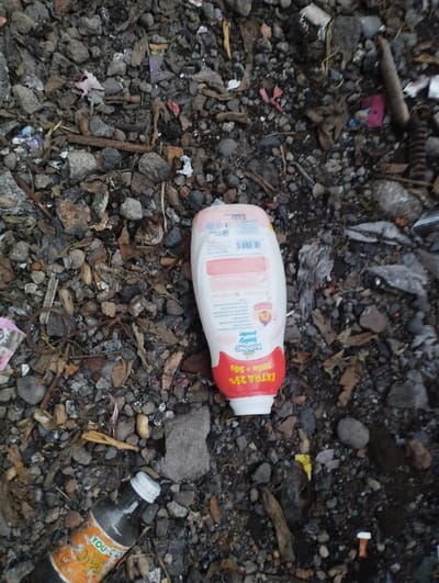 The image provides a top-down view of various discarded items lying on a rough, dirty, and gravelly ground surface in what appears to be an outdoor, unkempt area within Kecamatan Panji, Indonesia.
The central focus is a white plastic bottle with a red base and cap, positioned slightly off-center. This is identified as a "SoCoNo Baby Powder" bottle, with visible text on its red label stating "EXTRA 25% 200g + 50g." Additional smaller text, including "BPOM" (likely indicating Indonesian regulatory approval) and other product details, is visible on the bottle's white section.
To the bottom left, a clear glass bottle with an orange label and a white screw cap is lying horizontally. Part of the label clearly shows the brand name "YOU-C."
The ground is a mix of dark grey and brown pebbles, small rocks, and loose soil. Numerous pieces of litter are scattered throughout, including colorful plastic scraps (red, pink, purple), dried leaves, small twigs, bits of fabric, and other unidentifiable debris, contributing to an overall sense of disarray and pollution.
No people or activities are present in the image. The lighting is diffused, suggesting it is daytime, possibly under an overcast sky, as there are no harsh shadows. The scene conveys an impression of environmental neglect and the presence of discarded consumer goods.