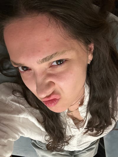 A close-up, high-angle image depicts a young adult female in an indoor setting within Vandœuvres, Switzerland. The subject, positioned with her head tilted, maintains direct eye contact with the camera. Her facial expression is characterized by subtly furrowed eyebrows and pursed lips. She has long, dark brown wavy hair, dark brown eyes, and fair skin with minor blemishes visible on her forehead. She is wearing a light-colored, possibly white, collared shirt with visible texture. Multiple thin necklaces with small pendants are layered around her neck, and a small silver hoop earring is present in her visible right earlobe. The background is out of focus, revealing a dark, monochromatic surface, likely fabric, indicating an indoor environment.