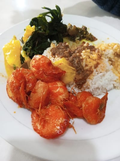 A white plate holds a meal featuring several fried shrimp with reddish-orange shells dominating the bottom left. Steamed white rice is topped with a dark brown sauce. Additional elements include vibrant green vegetables, a yellow-orange dish, and a small serving of a green and brown mixture. The dish is presented on a white surface, likely a table, with a small portion of blue fabric visible in the upper right corner.