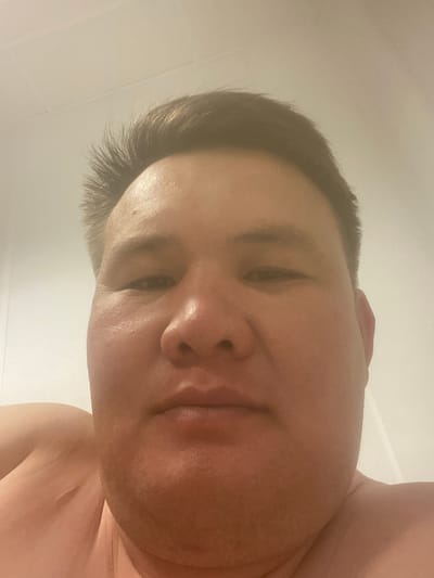 The image is a close-up, low-angle, selfie-style photograph of a person's face. The subject appears to be a man with a round face and short, dark hair. His skin is flushed, and there is visible pore texture. The background is a plain, light-colored wall with a subtle seam or line visible near the top of the frame. The lighting is soft and appears to be coming from above. The photograph captures the subject's forehead, eyes, nose, and mouth, as well as a portion of his shoulder and upper chest, which are bare. The location is identified as Қосшағыл ауылдық округі, Kazakhstan.