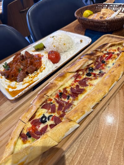 The image shows a table setting with food and drinks at a restaurant. In the foreground, there is a long, boat-shaped flatbread pizza topped with cheese, sliced salami, olives, and red bell peppers. This is served on a wooden board. Behind it, on a white rectangular plate, is a serving of rice, a roasted tomato, a green pepper, and what appears to be a dish of meat in a red sauce served over a white creamy sauce. In the background, a brown wicker basket contains bread and a lemon wedge. The table is wooden, and the chairs appear to be dark blue. The restaurant's ambiance suggests a casual dining experience. This scene is depicted at a restaurant located in Nottingham, United Kingdom.
