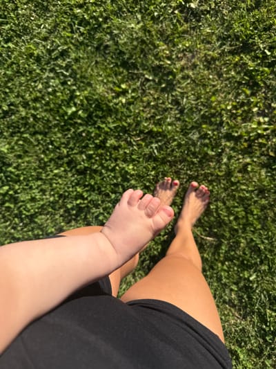 The image shows a top-down view of a grassy area, likely a lawn, with two pairs of feet visible. One pair of feet, belonging to an adult, has red nail polish. Adjacent to these, and slightly closer to the viewer, is a baby's foot and lower leg, which is lighter in skin tone and appears to be reaching out. The adult's legs are tan and wearing black shorts. The scene is outdoors, with natural lighting casting shadows on the grass. The location is identified as the village of Zazhevichsky Selsovet, Belarus.