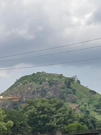 The image shows a lush, green hillside with rocky outcrops under a cloudy sky. In the foreground, dense trees obscure the lower portion of the hill. On the mid-level of the hill, there is a construction site with yellow machinery and scaffolding. Power lines are visible in the upper part of the image, stretching across the frame. The overall scene suggests a natural landscape with ongoing development.