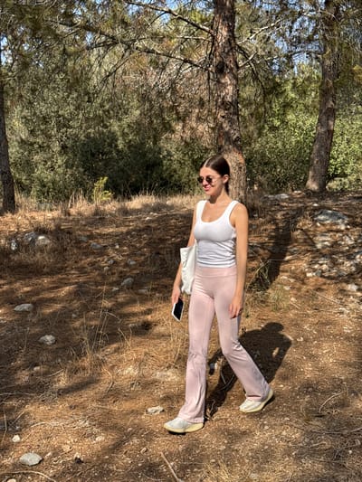 A young adult female is depicted walking outdoors in a natural, semi-forested area within or bordering the city of Modi'in-Maccabim-Re'ut, Israel. The setting features dry, earthy ground covered with brown pine needles, fallen leaves, small stones, and sparse dry vegetation, indicative of a sunny, warm climate. Multiple pine trees with rough trunks and green foliage are visible, casting distinct shadows on the ground, with denser green shrubbery in the background under a clear blue sky.

The subject, appearing to be in her 20s or early 30s, has dark hair tied back in a low ponytail and wears dark, round-framed sunglasses. She is dressed in a white tank top, light pink flared athletic pants, and light-colored athletic shoes. She is captured in a walking motion, stepping forward with her right leg, and her left arm swinging backward. She holds a black smartphone in her right hand and a white canvas tote bag is slung over her left shoulder. Her head is slightly tilted upward and to her left, revealing a smiling facial expression.