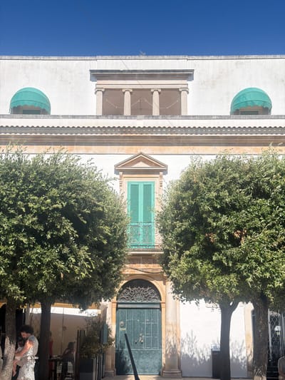 The image depicts a sunny street scene in Alberobello, Italy, focusing on the facade of a traditional building with a large, ornate green door and an arched entryway. Two lush green trees frame the entrance, their branches casting dappled shadows on the white wall behind them. On the left side, a man with curly hair is holding a baby. He is standing near the trunk of a tree. In the background, a woman is visible near a table, and another person is seated. Above the ground-floor entrance, a balcony with a turquoise shuttered window is visible, flanked by architectural elements that include columns and decorative awnings. The sky above is a clear, bright blue.