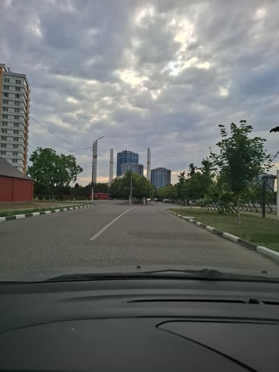 A street scene in Argun, Russia, is captured from the perspective of a vehicle's interior. The road, marked with white and black curbs, curves to the right. On the left, a tall, light-colored residential building with numerous windows dominates the foreground, next to a red brick building. Further down the street, a red bus is visible. In the background, several modern, multi-story buildings, including one with a distinctive blue facade, rise against a cloudy sky. Two tall, industrial-looking structures, possibly chimneys or cooling towers, stand between the buildings. Lush green trees line the right side of the street, some of which are young saplings in a manicured grassy area with a decorative fence. The lighting suggests it is either early morning or late evening, with the sky exhibiting patches of brightness breaking through the overcast clouds. A few individuals are sparsely scattered along the sidewalk.