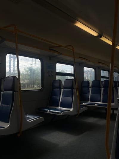 The image depicts the interior of a train carriage, likely in Aprelevka, Russia. The seats are upholstered in blue and white. Sunlight streams through the windows, casting shadows on the floor and illuminating the overhead fluorescent lights. Outside the windows, trees and power lines are visible. There are no people in the carriage, and the atmosphere is still and quiet, suggesting the train is either stopped or moving slowly. The overall impression is one of a common, functional public transport vehicle.