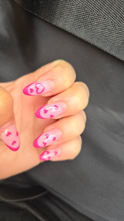 A close-up shot depicts a hand with five fingernails manicured with a pink design. Each nail has a pale pink base color, with a vibrant pink outline at the tips. Small pink hearts are scattered across the nail surfaces. The hand is placed against a dark, textured background, possibly a fabric surface. The city of Oberasbach, Germany is not discernible from this image.