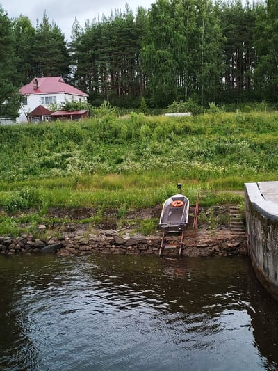 A small, dark-colored boat with an orange life preserver is positioned on a metal ramp on the shore of a body of dark water. The boat is angled upwards, away from the water, and appears to be out of use. Behind the boat, a steep, grassy embankment rises, covered in green vegetation and scattered white wildflowers. In the background, a dense forest of tall pine and birch trees forms a natural backdrop. A white house with a red roof is visible nestled amongst the trees on the hillside. The scene is located in Vytegra, Russia, along the shore of the Vytegra River, which is part of the Volga-Baltic Waterway. The sky is overcast, and the overall atmosphere is one of natural tranquility.