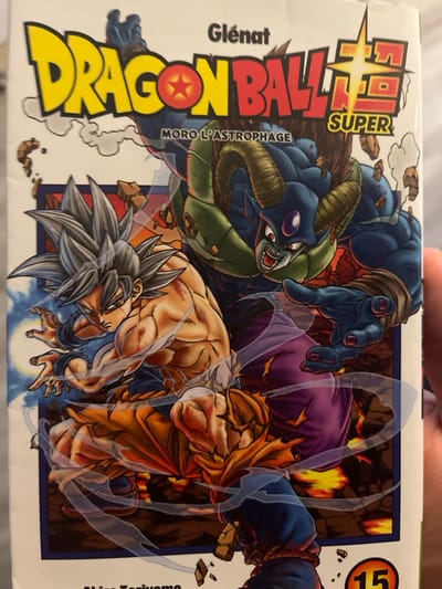 The image displays the front cover of a French edition of the "Dragon Ball Super" manga, volume 15, published by Glénat, subtitled "Moro L'Astrophage." The cover artwork features two characters in a dynamic, confrontational battle pose. On the left, a muscular individual with silver spiky hair, a determined expression, and a partially torn orange gi revealing a bare, scarred torso, is depicted in a fighting stance with blue energy effects around his forearms. On the right, a large, blue-skinned, horned character with green chest armor and purple pants, identified as Moro, reaches forward with an open hand and a menacing expression. The background illustrates a scene of fiery, rocky destruction with debris flying. The bottom right corner of the cover shows the number "15" within an orange circle. A human hand, with fingers visible on the right side, partially holds the manga volume. The artwork depicts a fictional battleground. The physical location where this photograph of the manga volume was taken, specifically within the city of Vieux, France, is not discernible from the visual evidence presented in the image.