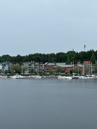 The image displays a waterfront scene in Lappeenranta, Finland, captured under an overcast sky that appears light blue-gray. The foreground consists of calm, rippled gray water. In the midground, a marina stretches along the shore, populated by numerous moored boats. These include various white pleasure crafts, both motorboats and sailboats with visible masts, interspersed with red buoys. A paved area or roadway borders the water, where multiple cars are parked.
The background features a blend of architecture and natural landscape. On the left, two modern, multi-story residential buildings with balconies and flat roofs exhibit facades of mixed light and reddish-brown tones. Adjacent to them on the right, a sequence of older, brick-built structures with sloped gray and red roofs, one featuring a prominent chimney, suggests former industrial or repurposed buildings. Behind these structures, a dense forest of predominantly green trees covers an elevated hill. Ascending above the treeline, two distinct tower structures are visible: a lattice telecommunication tower with multiple antennas and a smaller, auxiliary tower. No individuals are present within the captured scene.