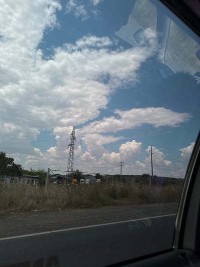 This is a vertical, eye-level, full shot of a power line tower and infrastructure viewed through a car window, with a reflection on the glass. The scene appears to be along a road in the city of Cherni Vrah, Bulgaria, on a partly cloudy day. The sky is a vivid blue with scattered white cumulus clouds, some of which are large and billowy, while others are thin and wispy. A tall, lattice-style power line tower is prominently positioned in the middle ground, with several power lines stretching across the frame. To the left of the tower, there are some dilapidated structures and fencing, with dry grass and vegetation in the foreground. In the distance, rolling hills can be seen. The car's interior, including the window frame and part of the dashboard, is visible in the foreground and along the right edge of the image, with reflections of the sky and clouds on the glass. There are no people visible in the scene.