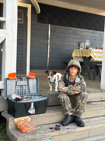 A young boy in camouflage clothing and black boots sits on a wooden deck next to a Jack Russell Terrier. The boy is wearing a hooded jacket, and his arms are crossed. The dog is looking towards the camera. A black open toolbox with orange latches is on the deck in front of them. To the right of the toolbox is a crushed orange bag of chips, and further to the right, a plastic water bottle. In the background, a dark gray wooden wall and a black shingled roof are visible. A striped yellow and white tablecloth is draped over a dark table. The scene appears to be outdoors, likely in a backyard or porch area. The lighting suggests it is daytime, possibly overcast. A sign on the door in the background has Cyrillic text that reads "ЕРЫХ". The overall mood seems relaxed and casual.