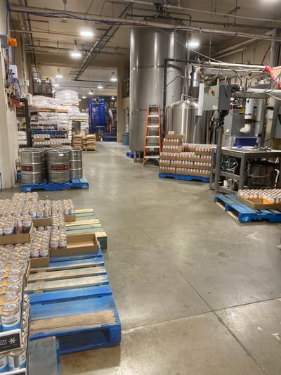 The image depicts the interior of a brewery or beverage production facility in Whitehorse, Canada. The environment is industrial, characterized by concrete flooring, exposed ceilings with piping and conduit, and large stainless steel tanks.
In the foreground, on the left side of the image, several pallets are stacked with cases of canned beverages. These cases are predominantly cardboard, and many of the cans are visible, displaying various colorful labels. Some of these cans are stacked in boxes, while others are arranged in rows on the pallets. One pallet in the immediate foreground has a portion of a blue wooden pallet visible.
Further into the scene, several large metal kegs, likely for beer or other beverages, are arranged on blue wooden pallets. These kegs appear to be made of stainless steel and bear some branding.
In the mid-ground, to the left, there are shelves stacked with bags of what appear to be raw ingredients, possibly grains or malt for brewing. There are also rows of smaller containers, potentially bottles or smaller cans, on shelves. A blue industrial machine is visible in the background behind these shelves.
Dominating the right side of the mid-ground and background are large stainless steel tanks, characteristic of brewing operations. Piping and various pieces of industrial equipment are connected to these tanks. An orange ladder is positioned near one of the tanks.
In the background, more industrial equipment and storage areas are visible, including more pallets of bagged materials and stacks of cardboard boxes. The overall lighting suggests an indoor industrial setting, likely during operational hours. There are no individuals clearly visible in the scene. The environment appears organized for production and storage.