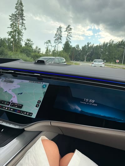 The image is taken from the interior of a car, looking out the windshield and at the car's dashboard displays. The car is in motion on a road, with trees and a cloudy sky visible outside. On the dashboard, a large screen displays a navigation map on the left side and the time and date on the right. The time displayed is 15:59, and the date is Tuesday, July 21, 2026. The location is identified as being in or near Ikšķile, Latvia.
