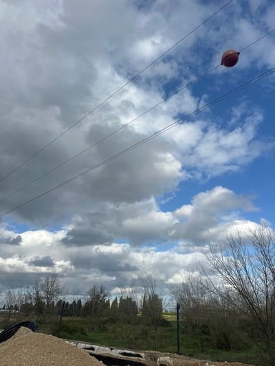 The image displays an outdoor scene in Kadima-Zoran, Israel, under a sky characterized by numerous grey cumulus clouds interspersed with patches of clear blue. Multiple parallel power lines extend diagonally across the upper portion of the frame. A spherical, reddish-brown aerial marker is attached to one of the upper power lines on the right side. The foreground features a pile of light-colored gravel and segments of black corrugated pipe, indicative of a construction or utility site. The midground contains a line of deciduous trees, currently bare of leaves, and taller evergreen trees. Beyond these trees, a field with green vegetation and a subtle hint of yellow is visible. No individuals, explicit actions, or interactions are present within the scene.