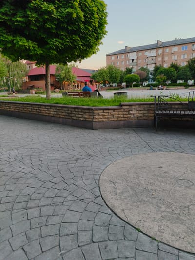 This media file depicts a public space in Bucha, Ukraine, likely a park or town square, during the late afternoon or early evening. The scene is dominated by a large, lush green tree in the foreground, with its canopy casting shadows on the patterned pavement. In the middle ground, a couple sits on a wooden bench, facing away from the camera, with the woman appearing to lean her head on the man's shoulder. Behind them, a building with a red roof is visible, possibly a cafe or restaurant, illuminated by string lights. Further back, a multi-story brick apartment building stands against a pale blue sky.
The pavement in the foreground is made of grey interlocking stones arranged in a circular, fan-like pattern. A raised brick planter with grass and a bench is situated between the pavement and the area where the couple is seated. The overall atmosphere seems peaceful and relaxed, with the soft lighting suggesting the end of a warm day. The presence of people enjoying a quiet moment in a public space, alongside urban architecture and greenery, creates a sense of everyday life. There is no visible text in the image.