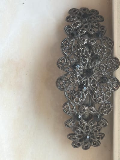 This is a close-up, vertical, eye-level shot of an ornate, dark metal hair clip adorned with black faceted gemstones. The hair clip is positioned on the right side of the frame, against a textured, light-colored background that appears to be paper or fabric. The background has a slightly mottled appearance with a few small, dark specks. The hair clip itself features intricate filigree work, forming floral or paisley-like patterns. The gemstones are embedded within these patterns, catching the light. The overall aesthetic suggests a vintage or antique piece of jewelry. There are no people or any discernible urban features of Arcahueja, Spain visible in this image.