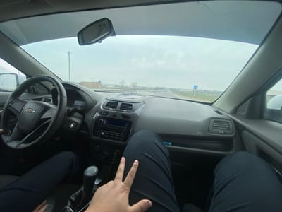 The image displays the interior of a Chevrolet vehicle, viewed from the front passenger seat looking forward. The driver's left hand is on the black steering wheel, and a manual gear shifter is visible in the center console. The front passenger's legs, dressed in dark trousers, and their right hand rest in the foreground. Through the windshield, a paved, two-lane road in Yettitom, Uzbekistan, stretches into the distance under an overcast, light gray sky. The surrounding environment consists of flat, open terrain with sparse, leafless trees and distant low-rise buildings. A blue rectangular road sign with white symbols is visible on the right side of the road in the mid-ground.