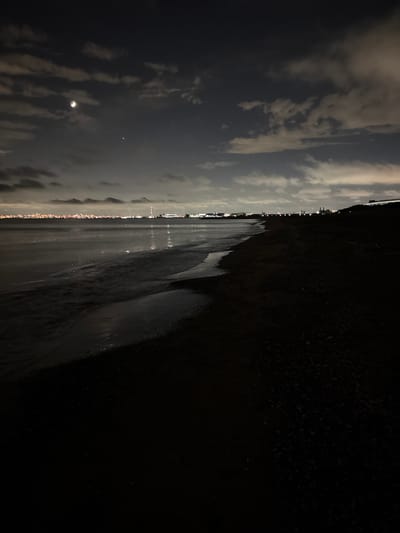 A nighttime view of a beach in Chiba, Japan. The moon is visible in the dark, cloudy sky, along with a bright star or planet. In the distance, a city skyline is illuminated with lights. The ocean water gently laps onto the dark sand of the beach. The scene is dark and moody, with the city lights reflecting on the water's surface.
