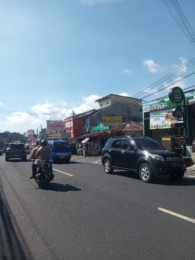 This image captures a street scene under a clear blue sky with scattered white clouds, situated in Ban Phonhxavanh, Laos. The perspective is from a road-level vantage point, looking along an asphalt multi-lane thoroughfare marked with a yellow dividing line.

In the immediate foreground, a black motorcycle with two individuals wearing helmets travels away from the camera. The rear rider wears a tan or light brown jacket. Behind them, a blue minivan with a license plate starting with 'D' is visible, also moving away. Further down the road on the left, a dark-colored SUV is present in traffic.

On the right side of the frame, a black Toyota Rush SUV with Indonesian license plate 'F 1048 YJ' is parked or moving slowly near the curb. Behind it, a commercial building features a prominent 'BURGER BANGOR' sign with a circular burger logo and additional text 'BUKA 24 JAM' (Open 24 Hours in Indonesian). To the left of this, another building has a red facade, with a partially visible sign resembling 'INDOMARET' and also displaying 'BUKA 24 JAM'. A green sign above it reads 'SINAR RAYA'. Multiple utility lines traverse the upper portion of the scene. Strong shadows cast by vehicles on the road indicate midday conditions.