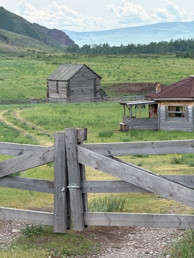 The image shows a wooden fence in the foreground, with a gate secured by a metal latch. Beyond the fence, a grassy field stretches out, with a dirt path winding through it. In the mid-ground, there are two rustic wooden structures. The one on the left is a log cabin, and the one on the right appears to be a small house with a porch and a red corrugated metal roof. In the background, a wooded area can be seen, followed by rolling hills and mountains under a cloudy sky. The scene is set in a rural, natural environment, possibly the village of Anchyl-Chon, Russia, characterized by its remote and pastoral landscape.