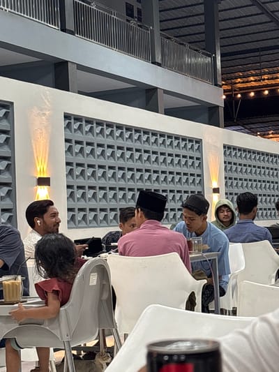 This image captures an indoor or semi-open dining area, likely a cafe or restaurant, in Kampongbaro, Indonesia. The setting features a modern industrial aesthetic with a corrugated metal roof, exposed dark structural beams, and white walls. A prominent design element is the decorative concrete block pattern on the upper part of the walls, illuminated by warm-toned wall sconces.

Several people, predominantly men and one young child, are seated at tables with white plastic chairs. The general atmosphere appears relaxed. Most of the men are dressed casually; one wears a black peci (a traditional cap), another a black kufi, and one is seen in a light-colored hoodie. One man has wireless earbuds in his ears. They are looking down, possibly at phones, or engaging in quiet conversation. A glass containing a reddish beverage, likely tea, is visible on one of the tables. A young child, with dark curly hair and wearing a red top, is seated in a gray high chair, looking down.

The warm glow from the wall sconces and string lights on the roof structure suggests the photograph was taken in the evening or at night. There is some unreadable small text or labels on the back of the child's high chair. The scene conveys a sense of a casual gathering or meal amongst friends or family.