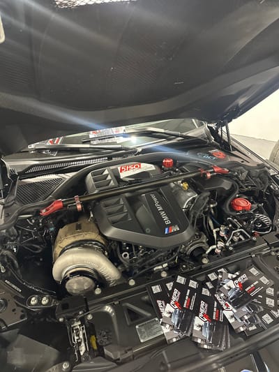 The image displays the open engine bay of a modified black BMW performance vehicle, situated within an indoor workshop or garage in Wolica, Poland. The underside of the raised hood reveals its carbon fiber construction. A prominent black engine cover with "BMW M Power" and the BMW M logo is central. To the left, a large, light-colored aftermarket turbocharger, partially encased in a beige heat-resistant wrap, is visible. A carbon fiber strut bar with red anodized adjustable end links spans across the engine bay, with "5150 Auto Sport" branding on one bracket. A red "5150 Auto Sport" sticker is present near the windshield wipers, and several components, including fluid reservoir caps, feature red accents. Multiple "CHEPA RACING" logos are visible, including one on a metallic plaque on the front crossmember. A fan of "CHEPA RACING SERVICE AND TUNING" business cards, featuring a car graphic and QR codes, is spread across the front of the engine bay. The scene is static, devoid of individuals or active work, illuminated by overhead artificial lighting reflecting off surfaces.