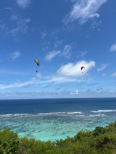 The image shows a coastal scene in Benoa, Indonesia. The sky is blue with scattered white clouds. Two paragliders are visible in the sky, one is red and the other is yellow with a dark colored individual hanging below. The ocean is two-toned, with deep blue water transitioning to lighter turquoise near the shore. Waves are breaking along a coral reef. The right side of the image shows a green lush coastline.